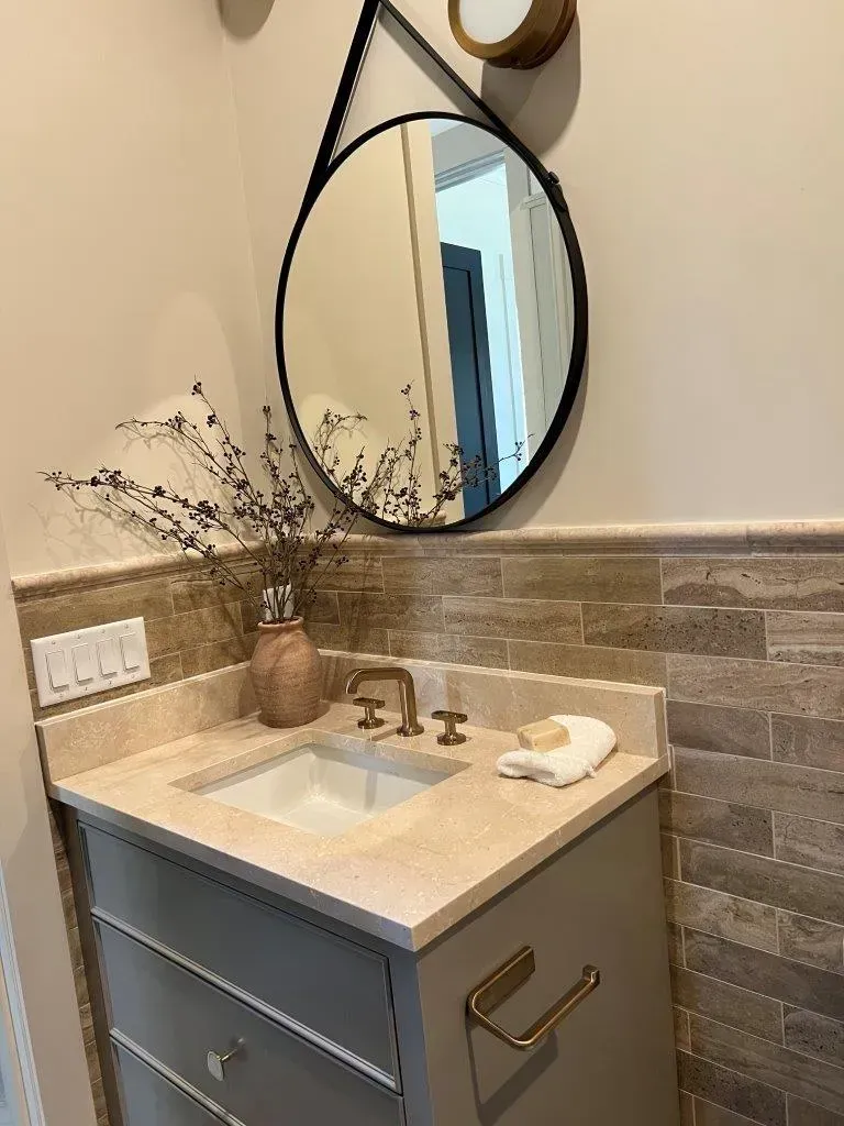 Small bathroom vanity with marble sink, round mirror, gold fixtures, and beige tile backsplash.