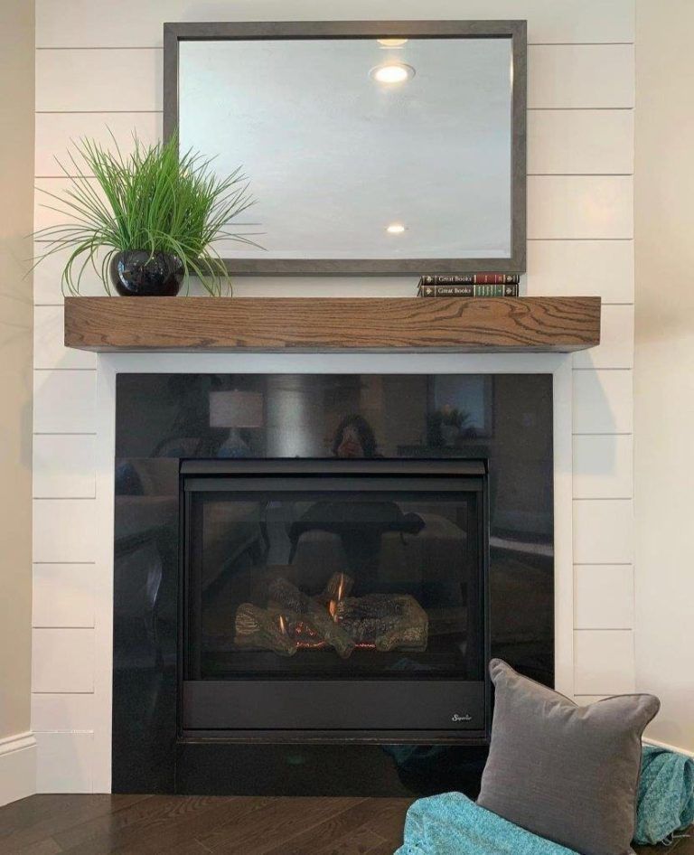 Modern fireplace with black insert, wood mantel, mirror, potted plant, and gray pillow in a bright room