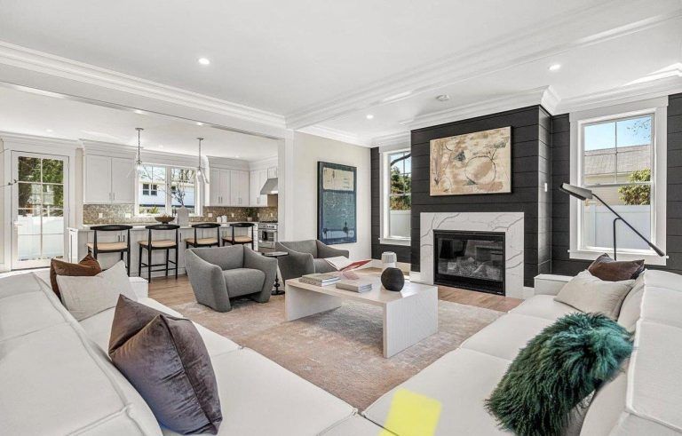Bright open living room with white sofas, gray chairs, a dark fireplace, and an adjoining kitchen.