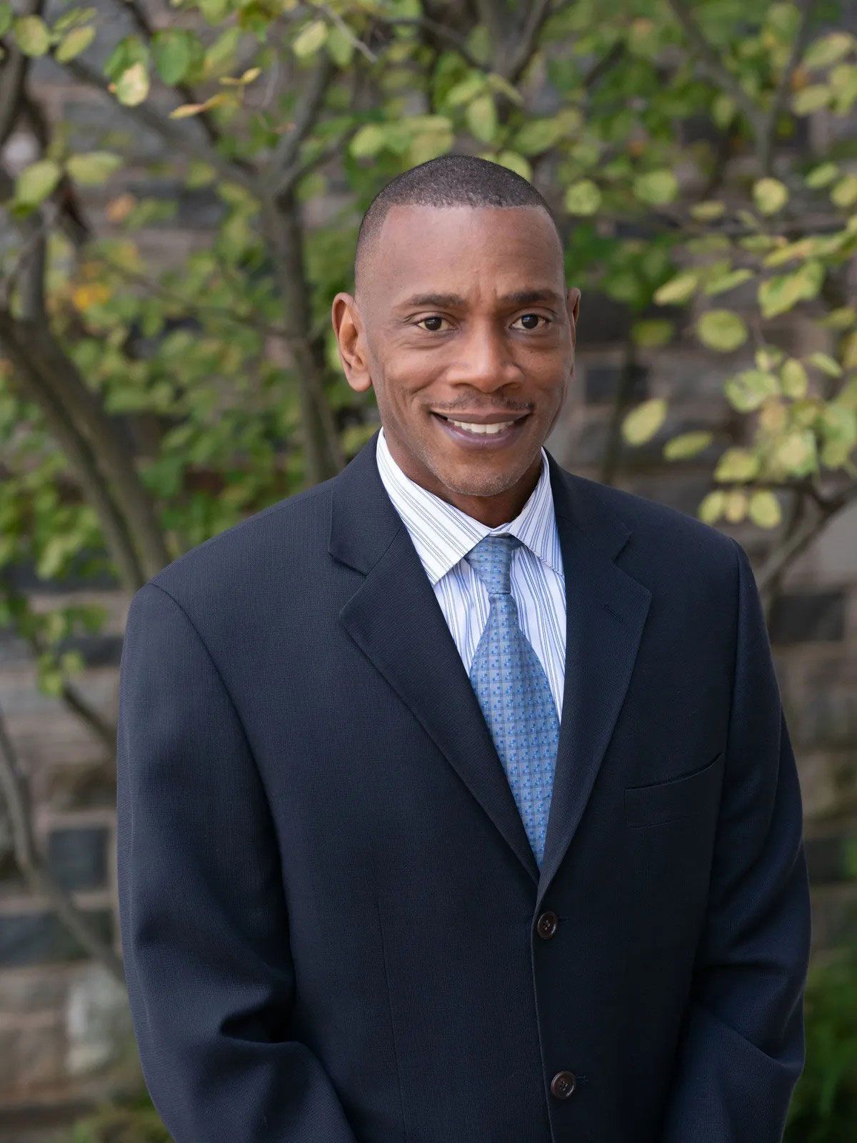 Man in a suit smiles outdoors, a blue tie, light blue and white striped shirt, and foliage background.
