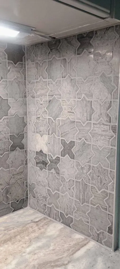 Gray tile wall with patterned design in a corner of a room, a small portion of the floor is visible.