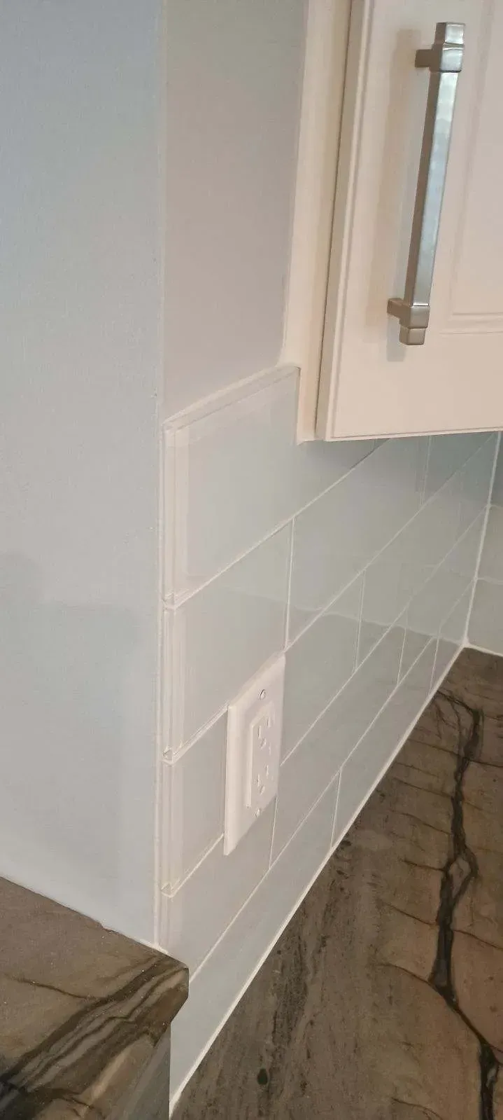 Corner of a kitchen backsplash with light blue rectangular tiles, an electrical outlet, and a cabinet.