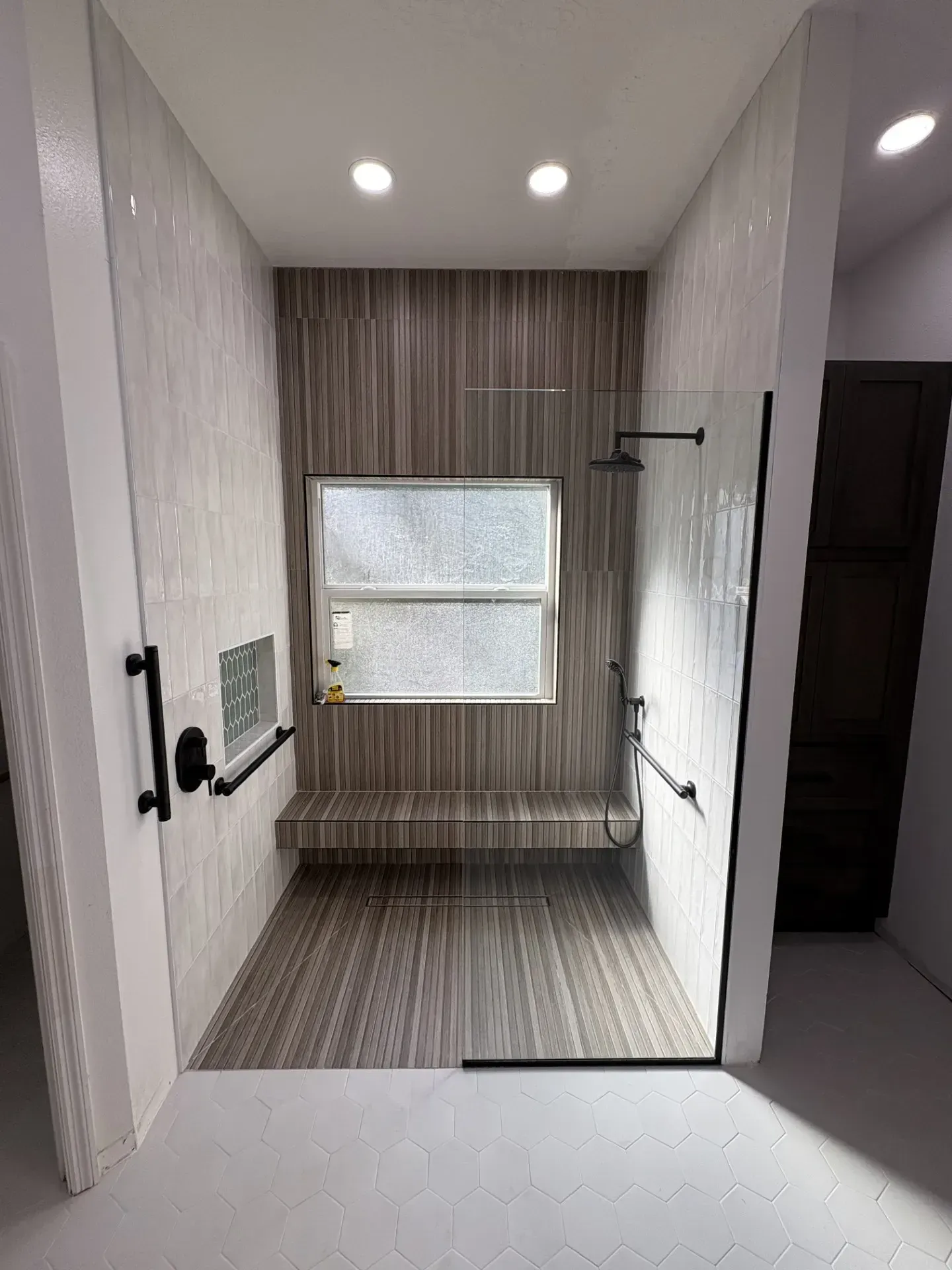 Accessible shower with glass door, bench, grab bars, and window. Beige and brown tones.