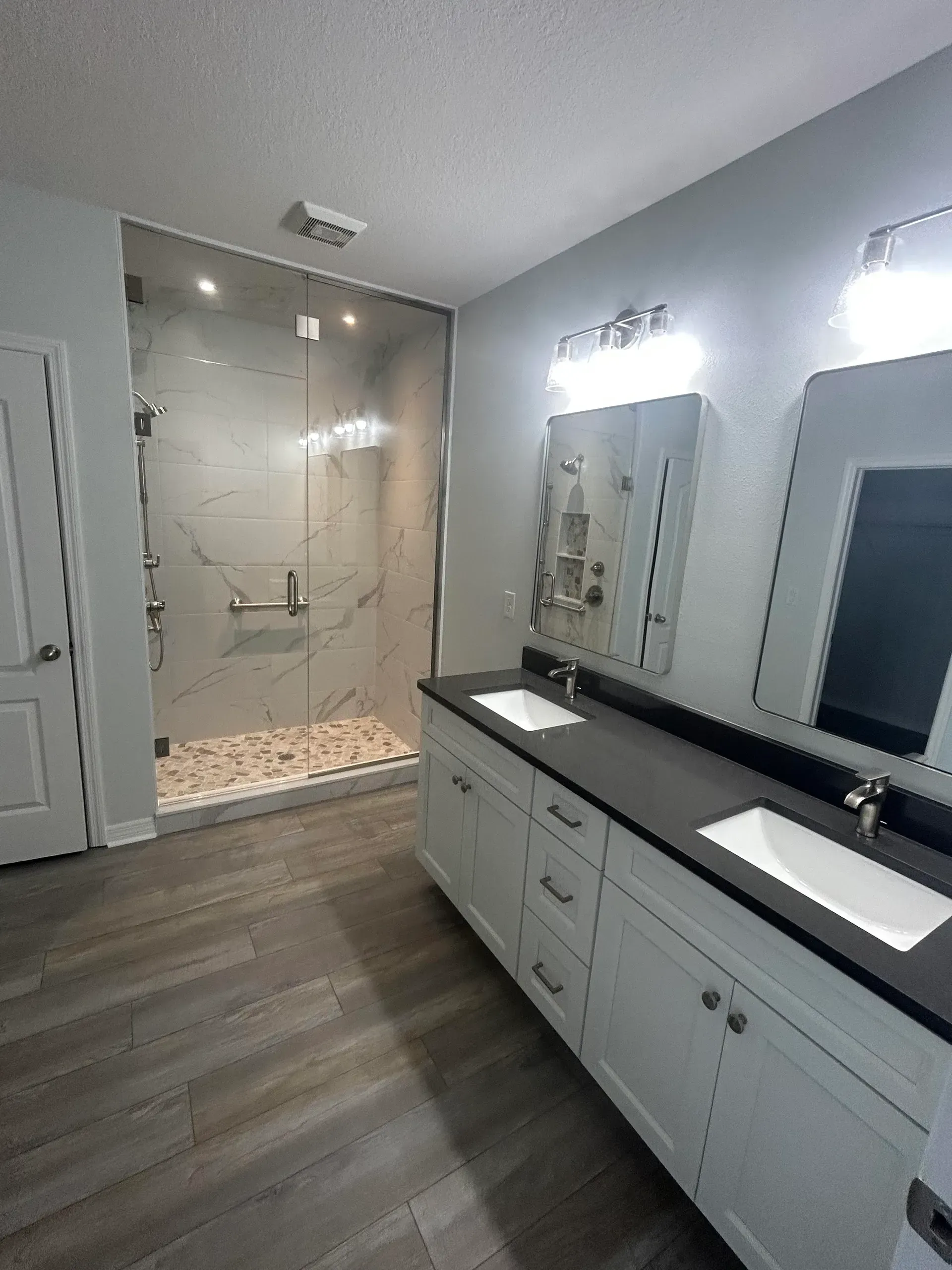 Bathroom with double vanity, shower, and gray flooring. White cabinets, black countertops, and glass shower door.