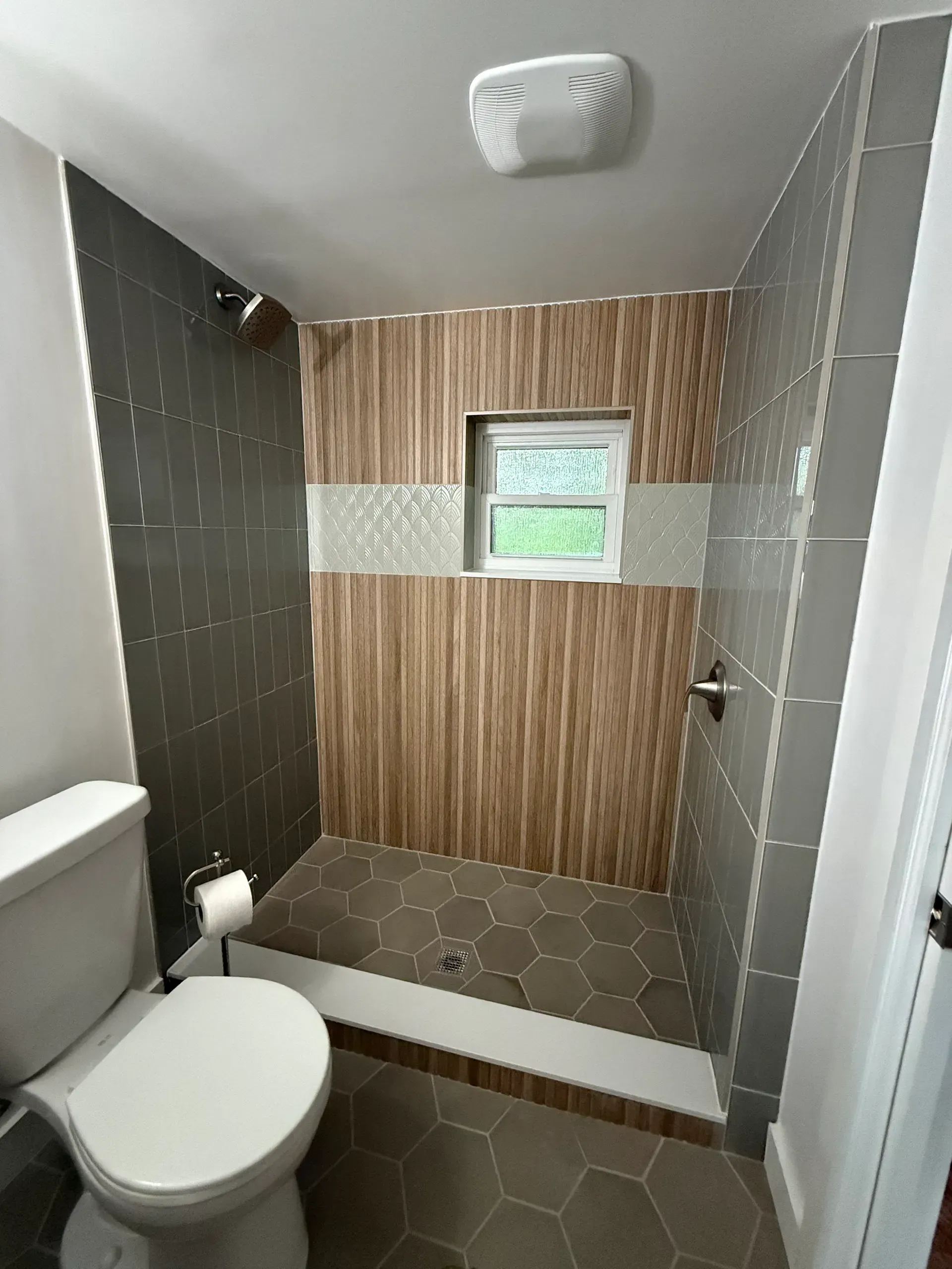 Bathroom with gray and wood-look tiled shower, toilet, and small window.