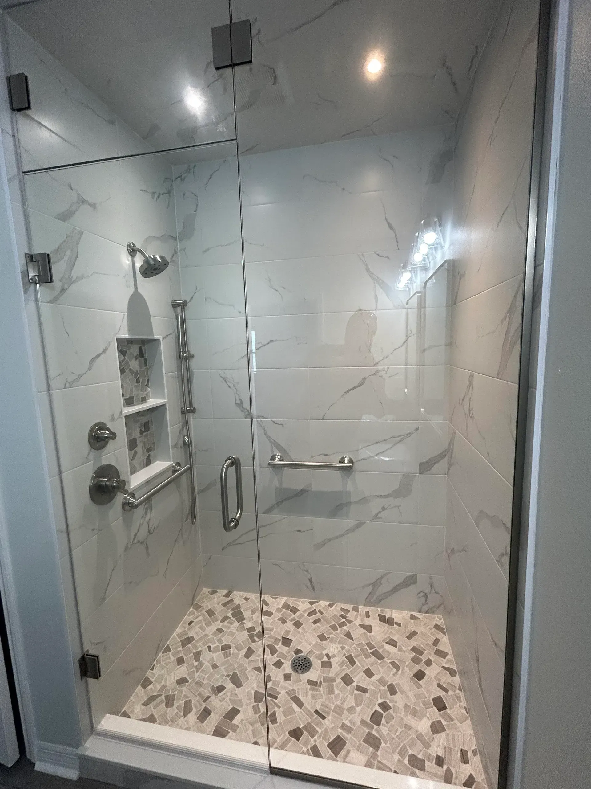 Glass-enclosed shower with white marble-look tiles, mosaic floor, built-in shelves, and grab bar.