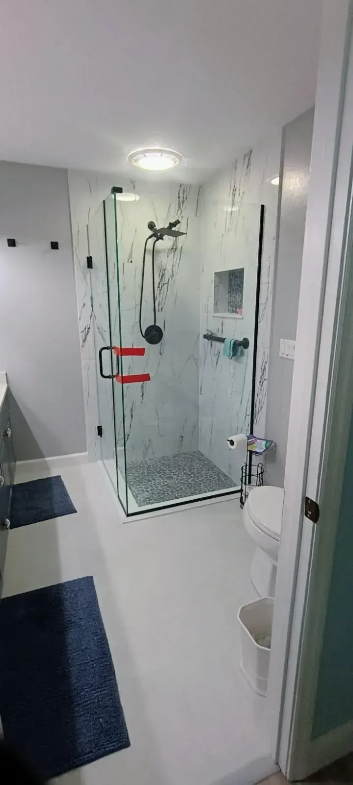 Bathroom with a glass shower, toilet, and blue rugs. White walls, gray walls, and red handles.