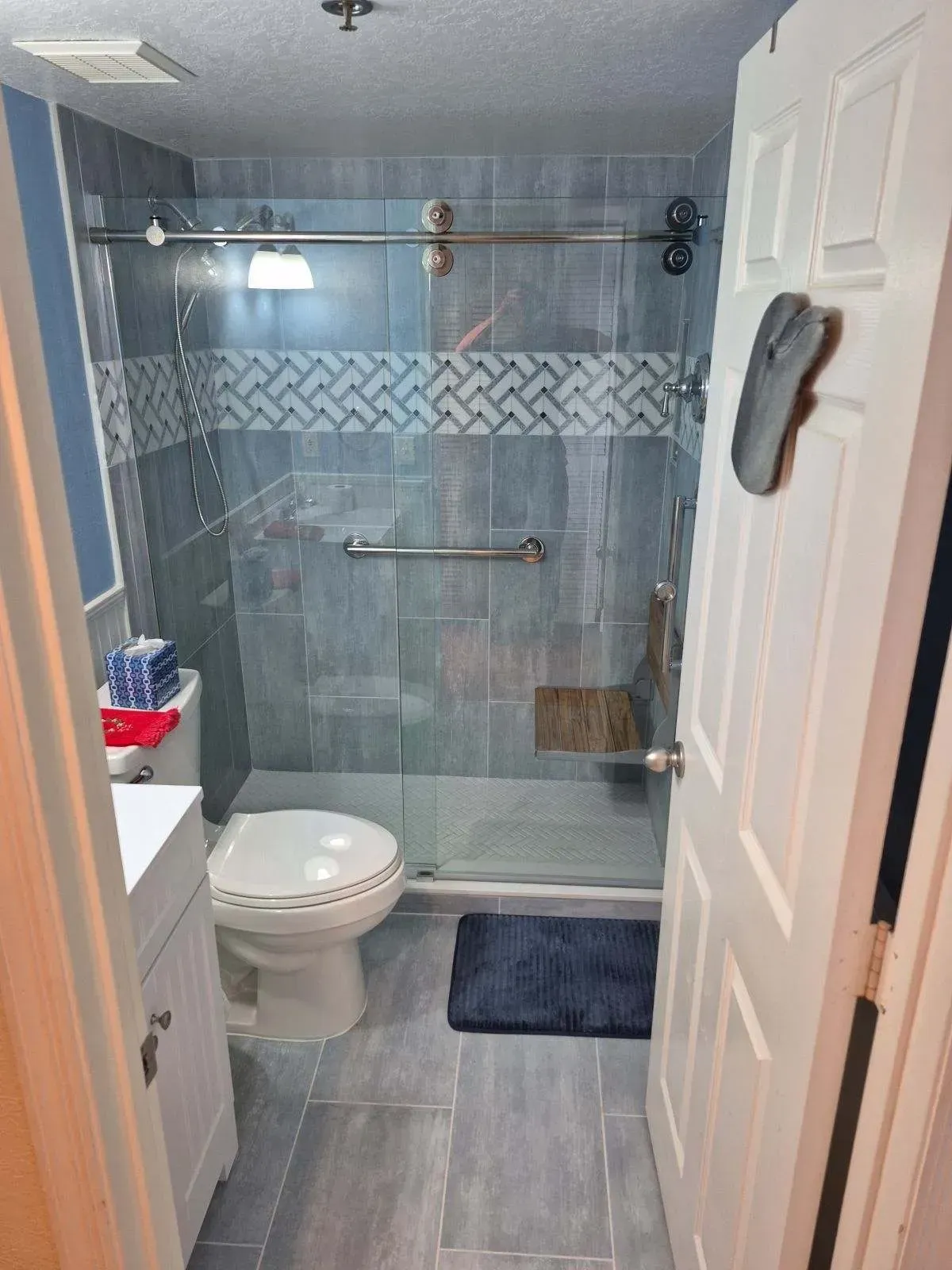 Bathroom with blue tile shower, toilet, and white door. Blue rug on floor.