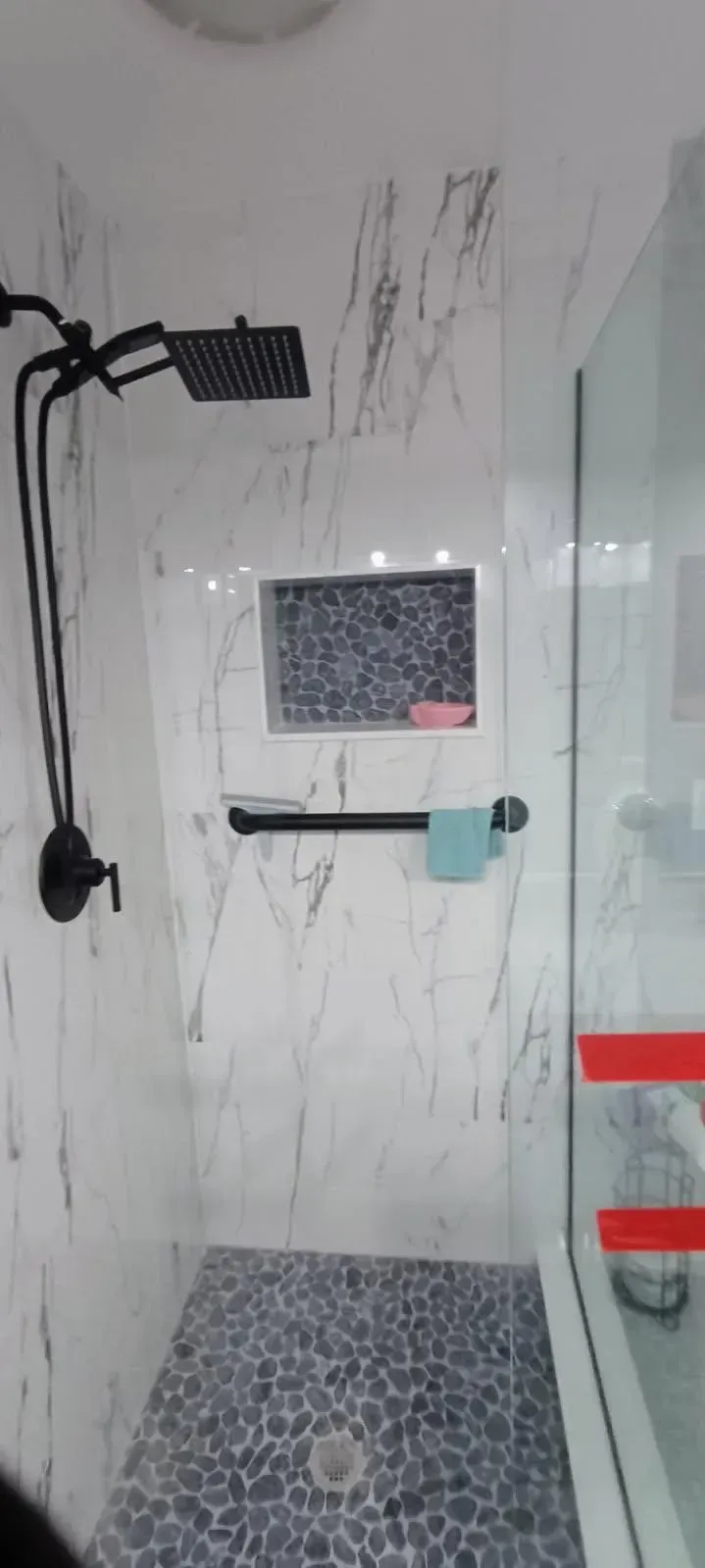 Bathroom shower with black fixtures, marble-look walls, pebbled floor, and glass door.