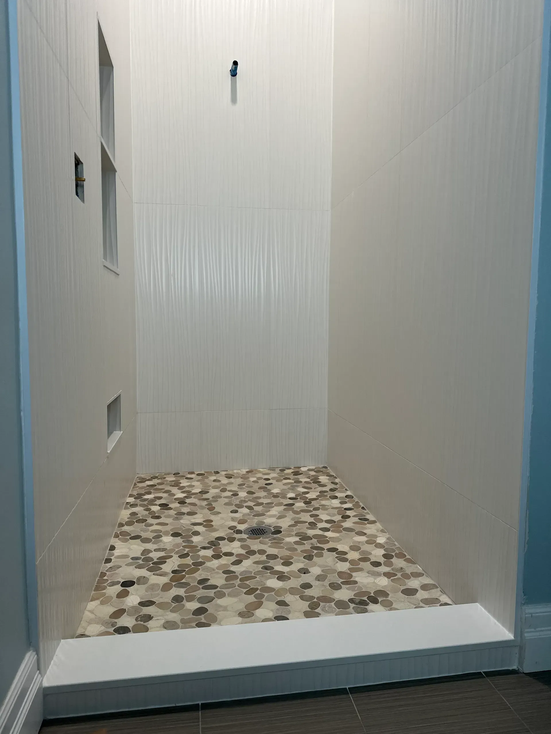 Empty shower stall with pebble tile floor and white textured tile walls.