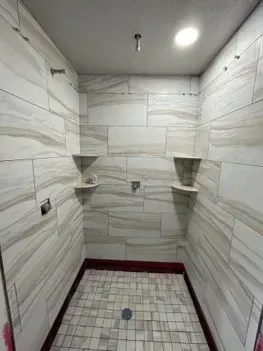 Shower stall with white and grey tiled walls, two shelves, shower fixtures, and a tiled floor.