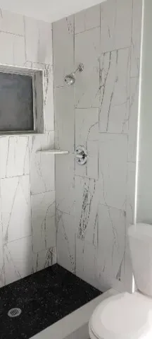 Shower with marble-like tile walls, black tile floor, and toilet in a bathroom.