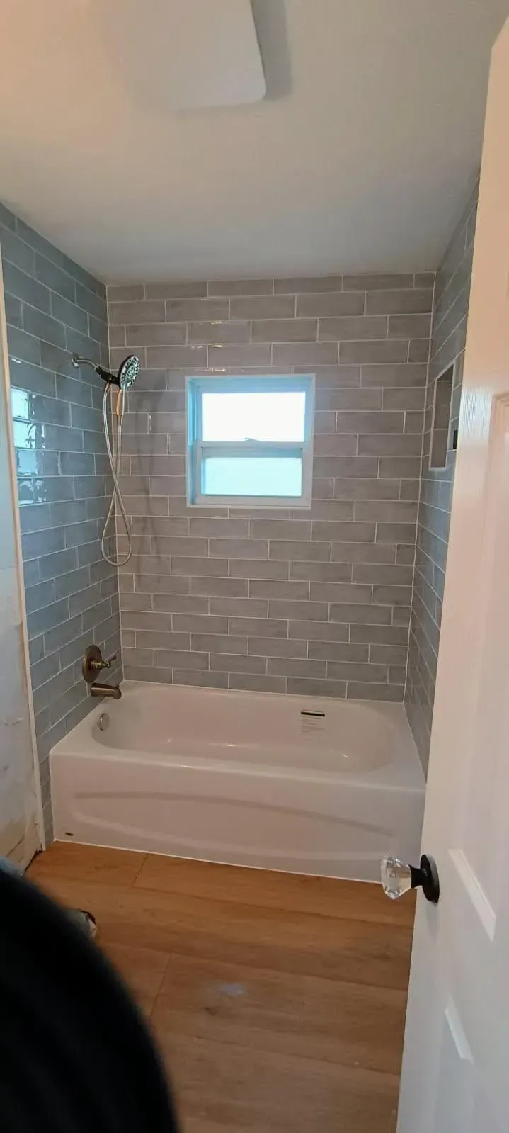 A bathroom with gray tiled walls, a white bathtub, and wood flooring. A window is in the wall.
