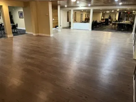 Spacious room with a polished hardwood floor. Tables and chairs visible in the background. Beige walls and recessed lighting.