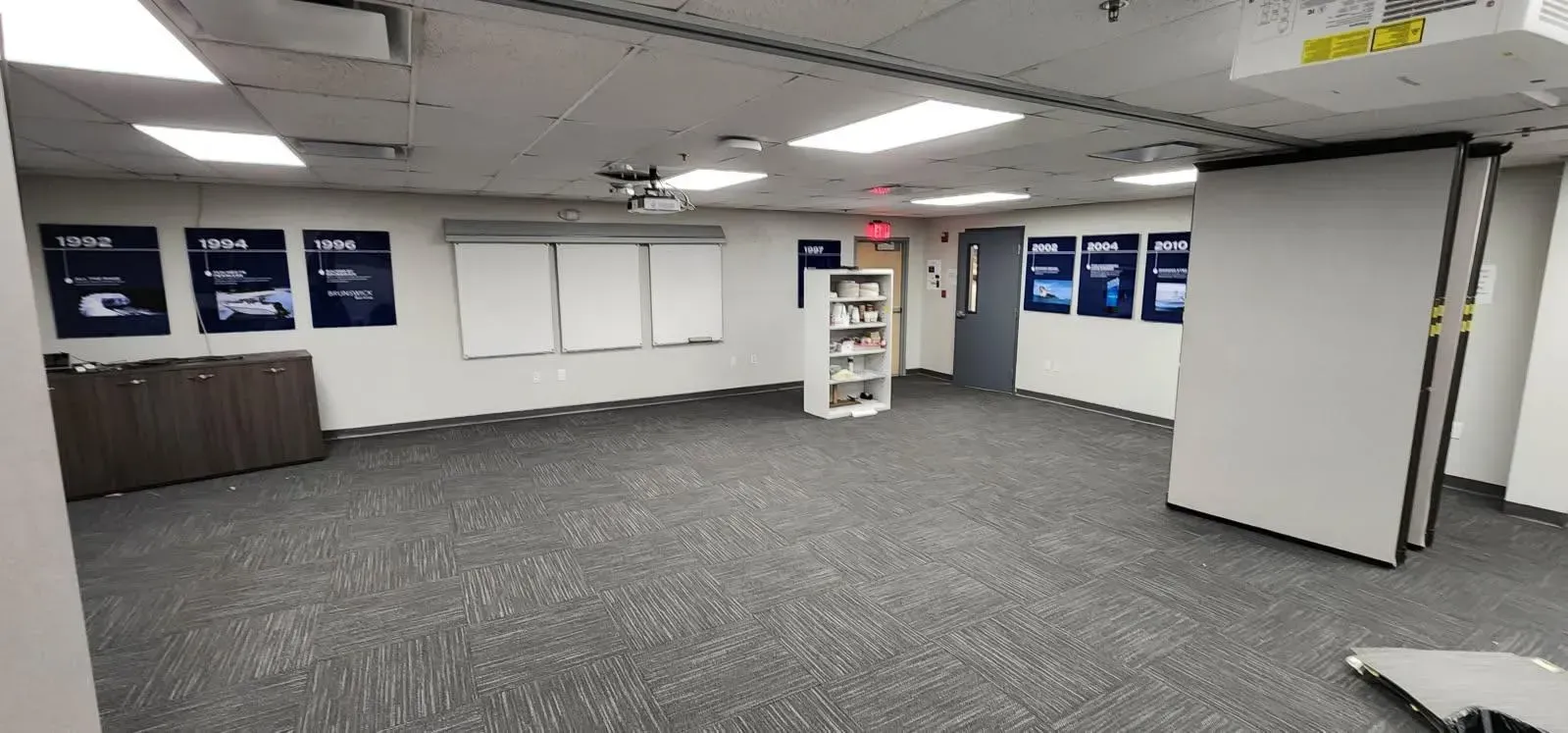 An empty office room with posters, whiteboards, and a bookshelf. The floor is carpeted.