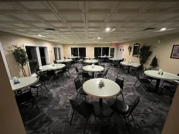 Empty restaurant dining room with round tables, black chairs, and windows.