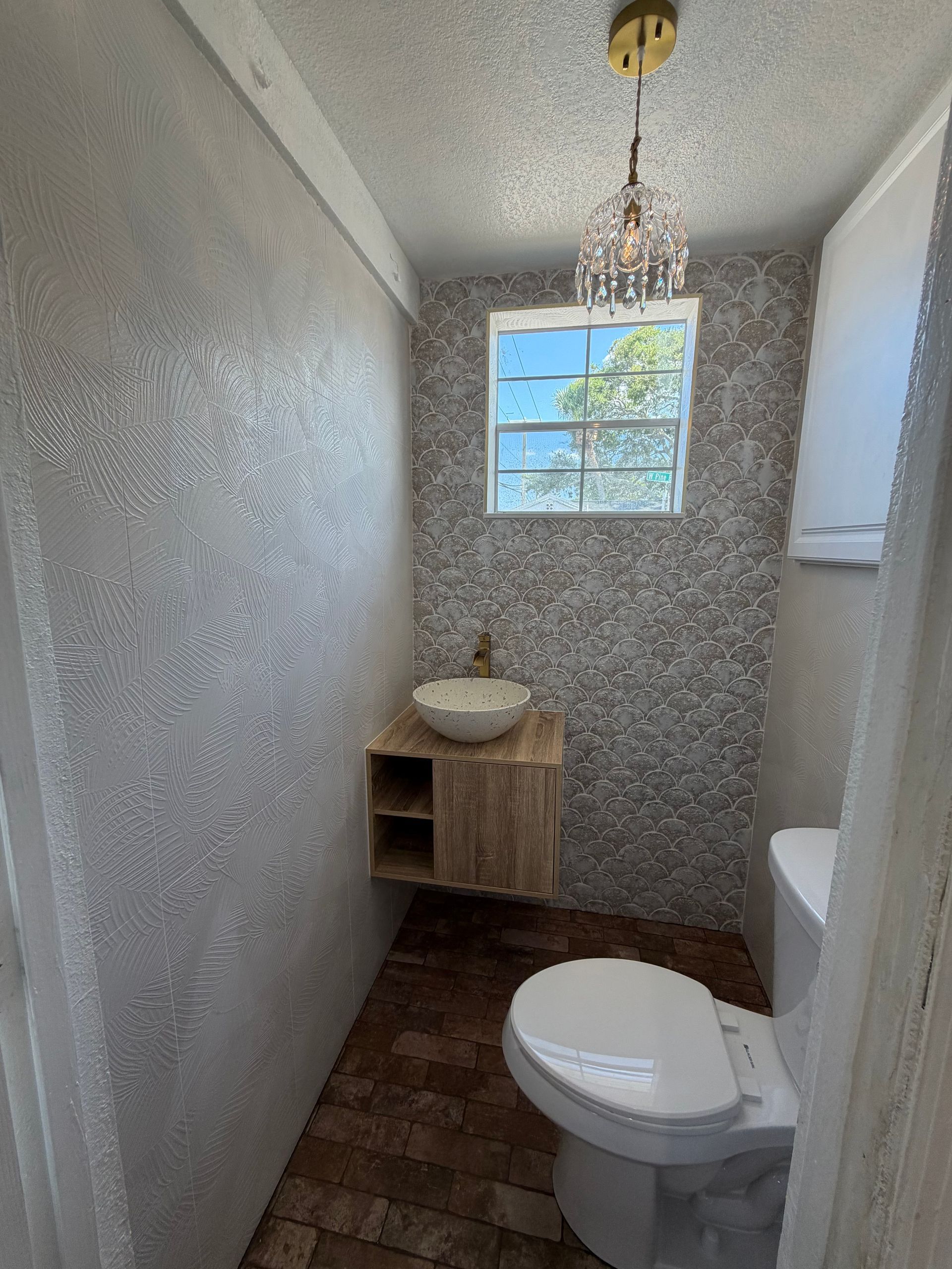 Narrow bathroom with textured gray walls, toilet, small sink, window, and hanging light fixture.