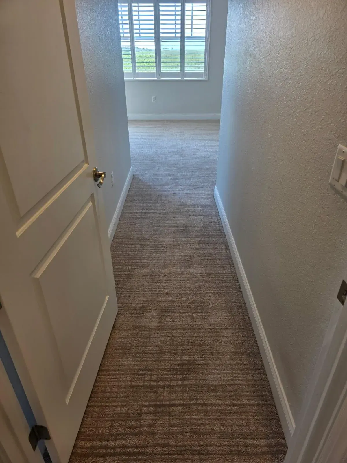 Narrow hallway with beige carpet leading to a bright room with window and white walls.