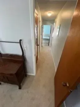 Hallway with light gray walls, carpet, and a wooden chest. A partially open wooden door is on the right.