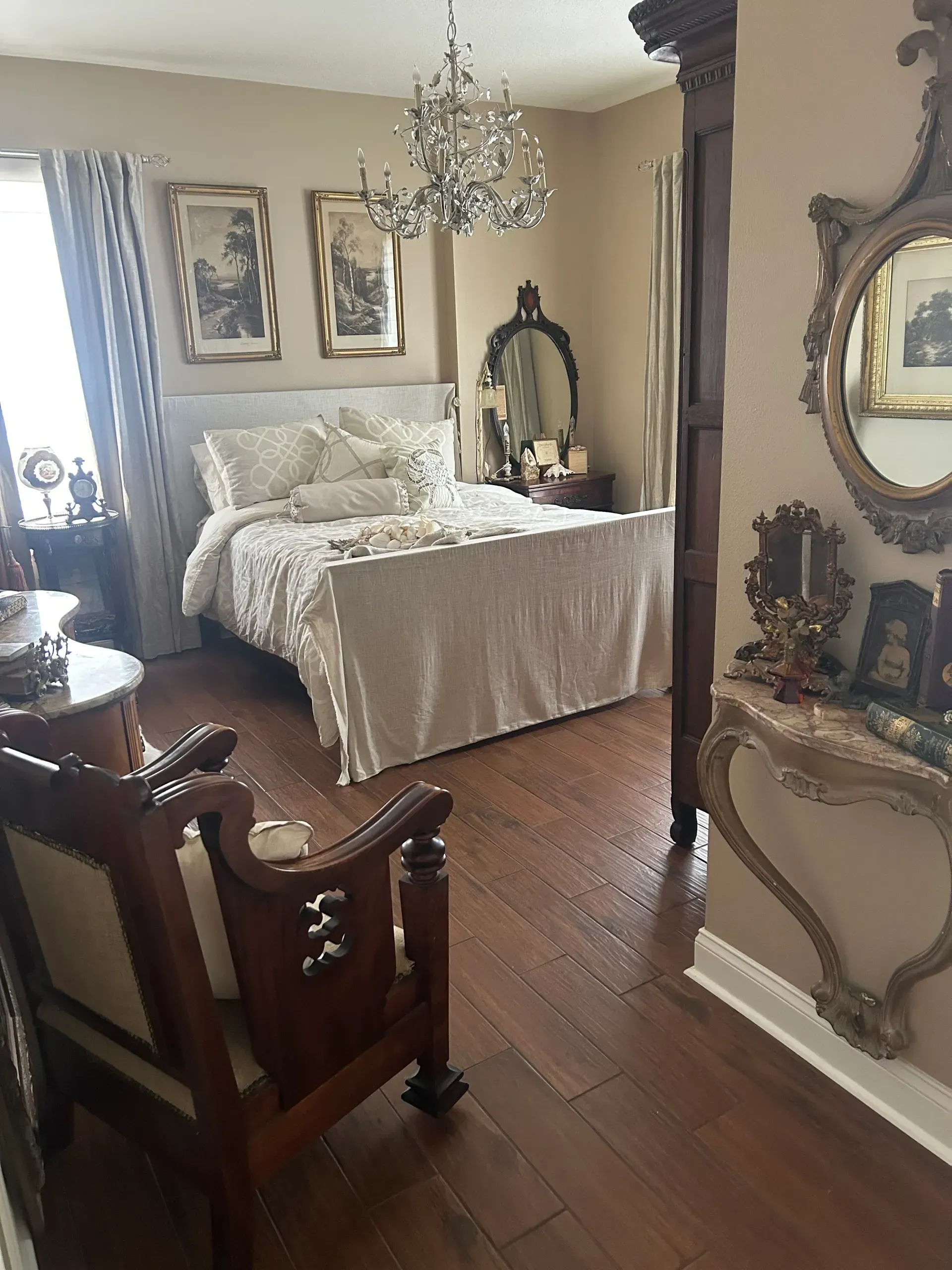 Ornate bedroom with chandelier, bed, antique furniture, art, and mirror. Hardwood floor.