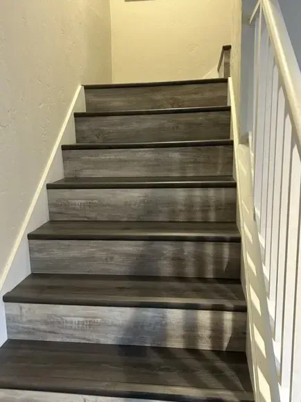 Wooden-look gray stairs with dark trim ascend toward a white wall. White handrail on the right.