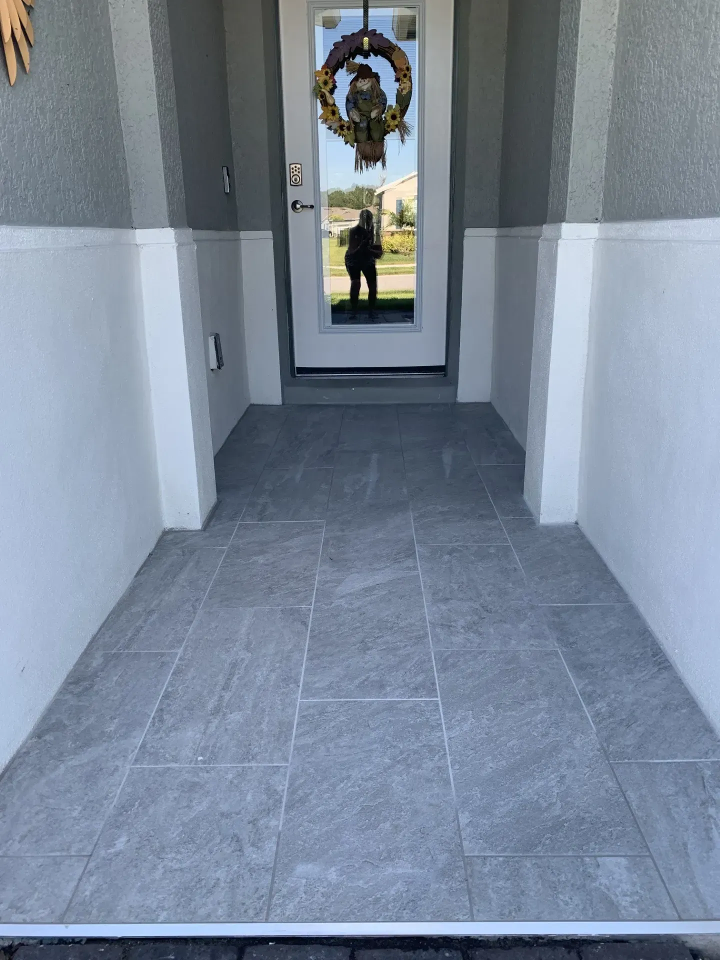 Gray tiled porch leading to a white door with a wreath. White walls surround the porch.