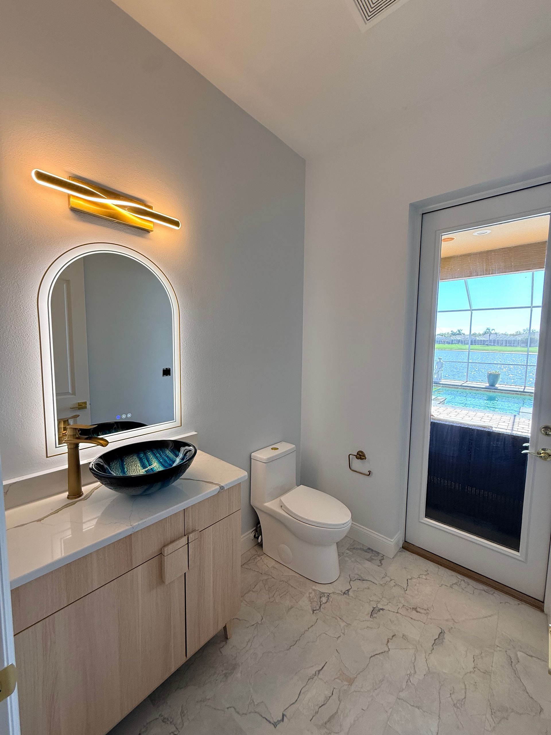 A modern bathroom with light wood vanity, arched mirror, dark vessel sink, toilet, and glass door overlooking a pool.