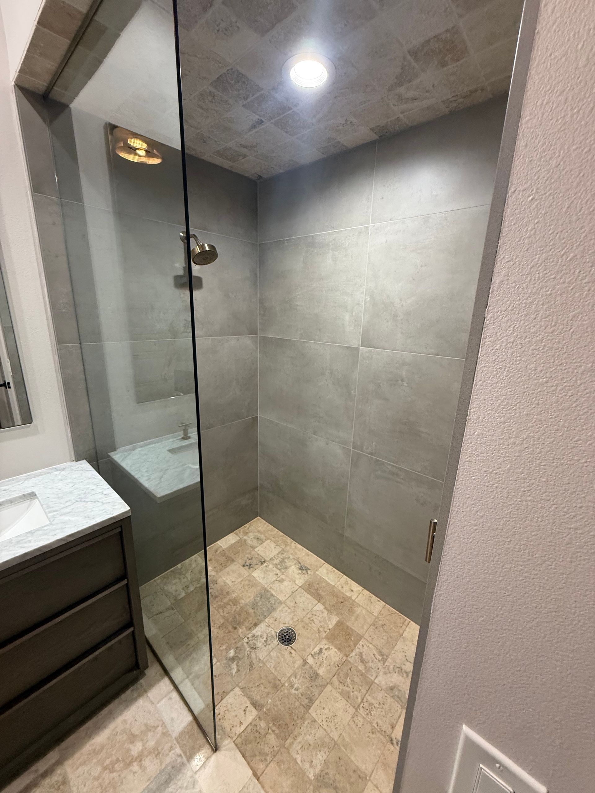Modern bathroom shower with gray tile walls, glass door, and beige tile floor beside a vanity.