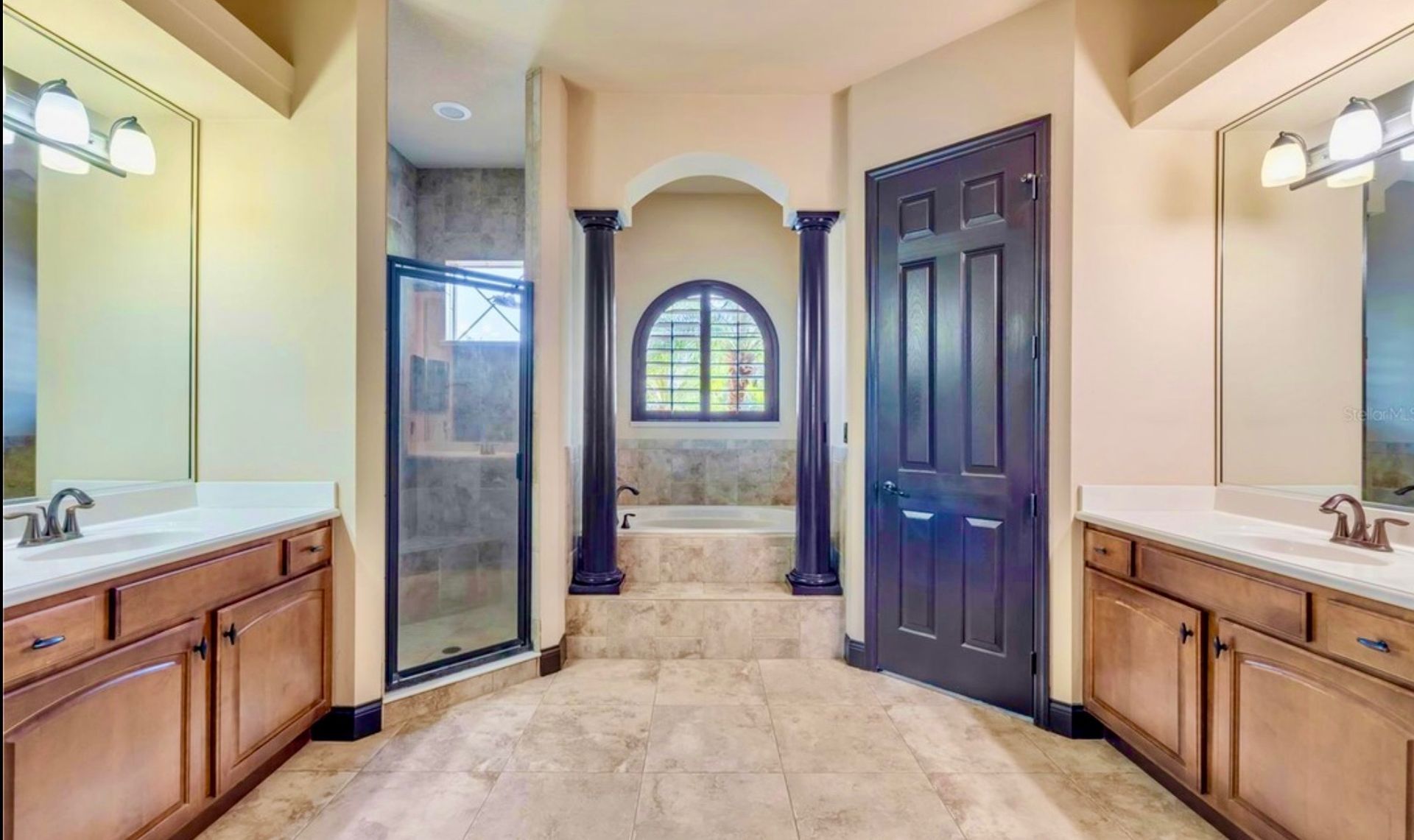 A bathroom with dual wooden vanities, a glass-enclosed shower, and a raised soaking tub area framed by two dark pillars.