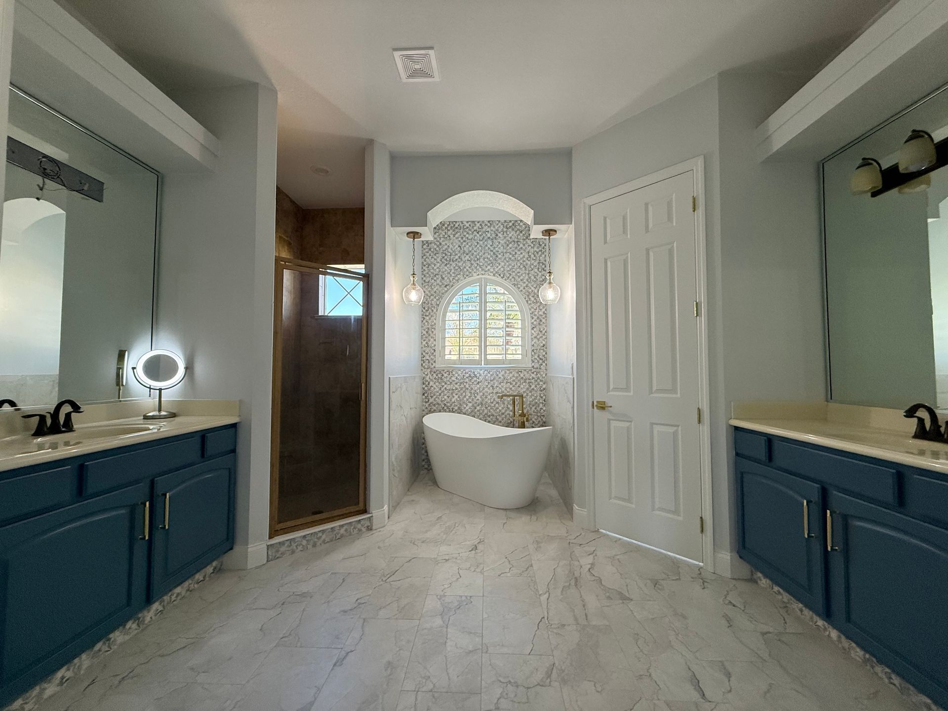 A bathroom with blue double vanities, marble-look tile floors, a white soaking tub under an arched tiled wall, and shower.