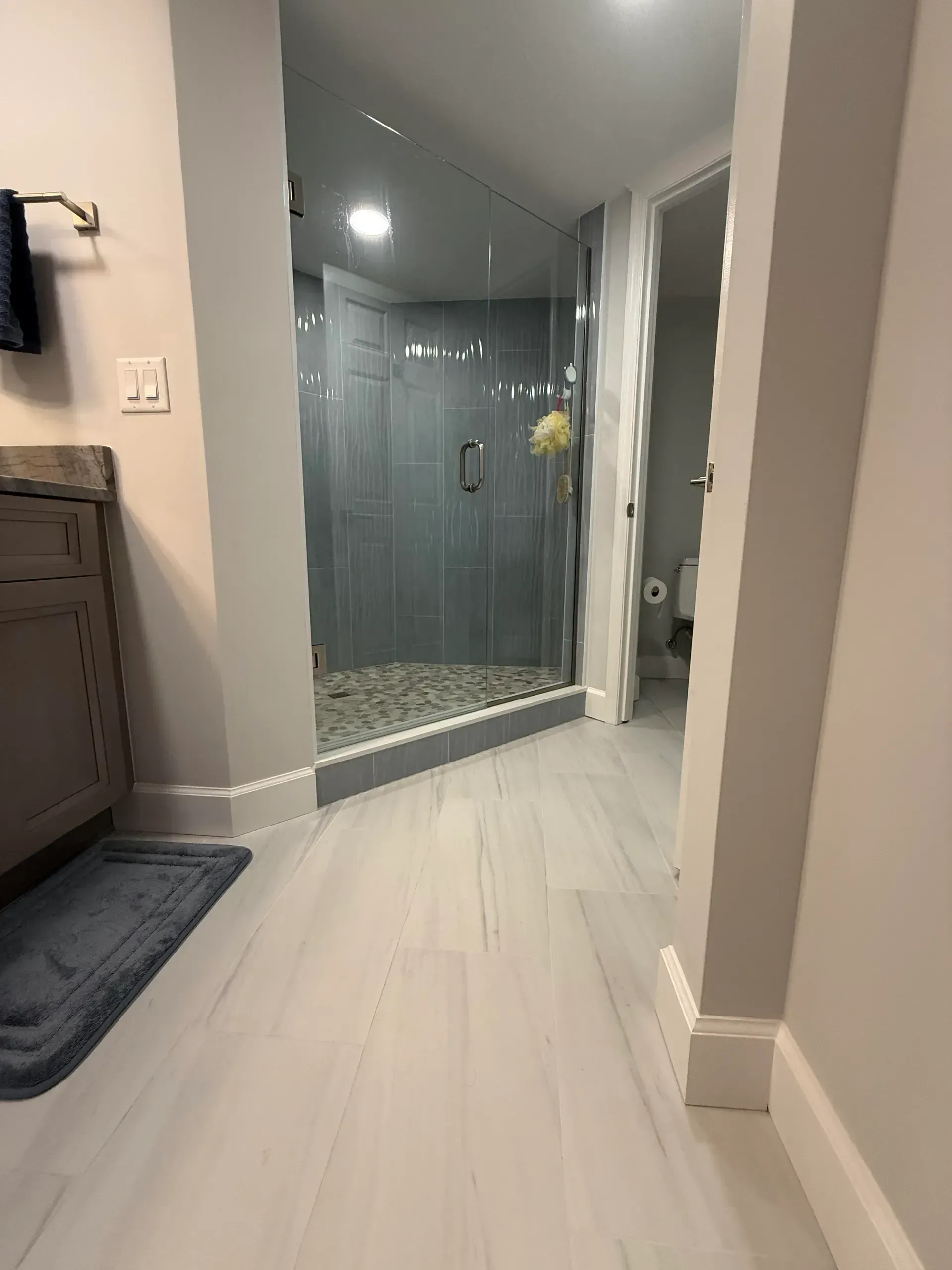 Modern bathroom with glass shower, white tile floor, and gray vanity seen from the hallway