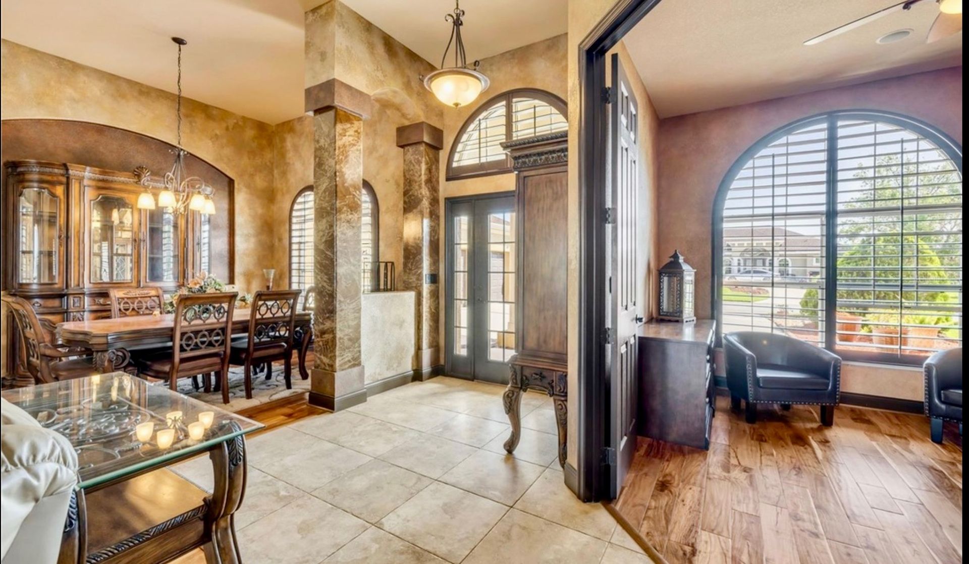 An elegant room featuring a dining set, marble tile floors, stone columns, and an archway leading to a sunlit seating area.