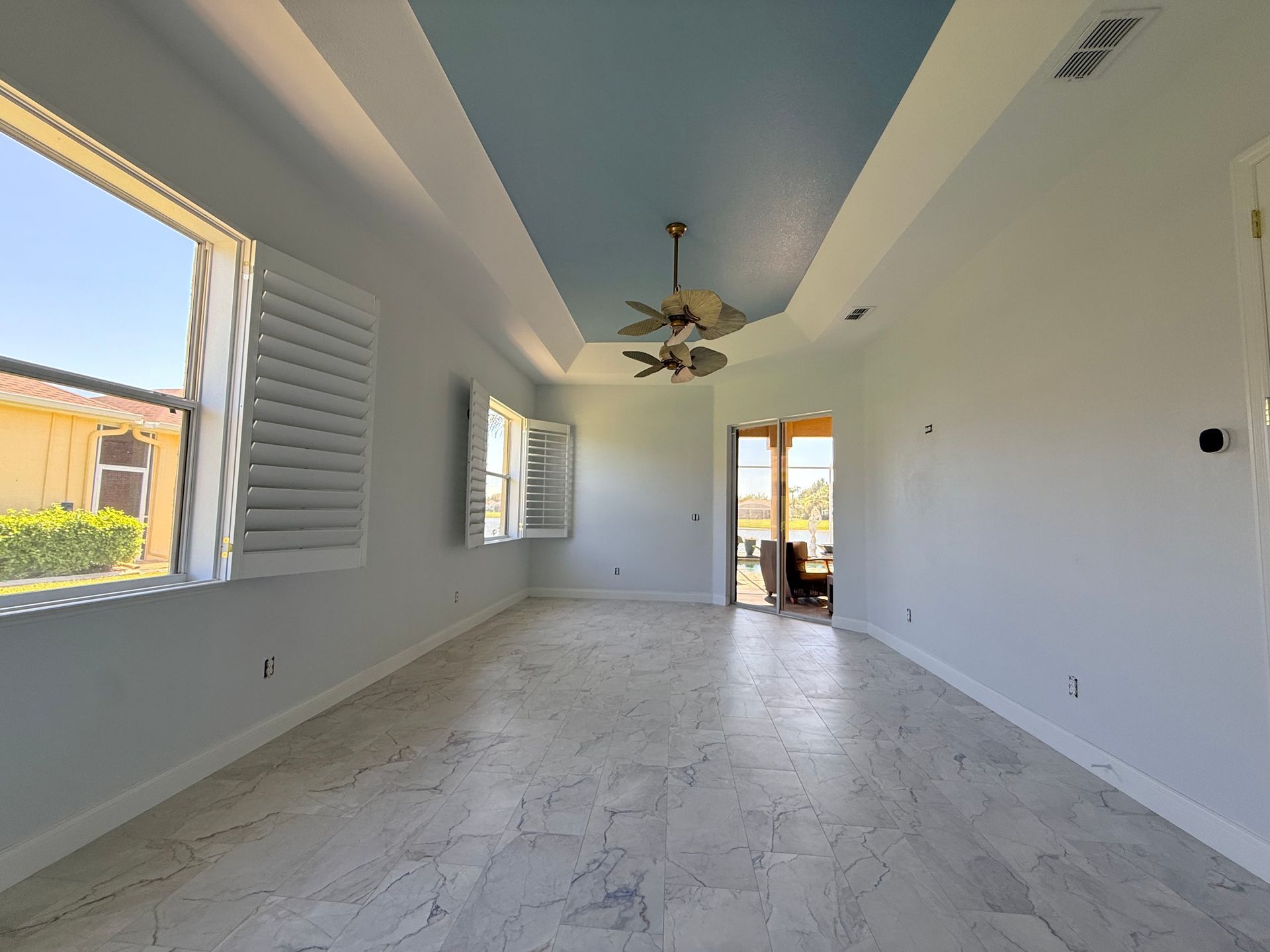 A spacious, sunlit room with white walls, a light blue tray ceiling, marble-look tile floors, and large window shutters.