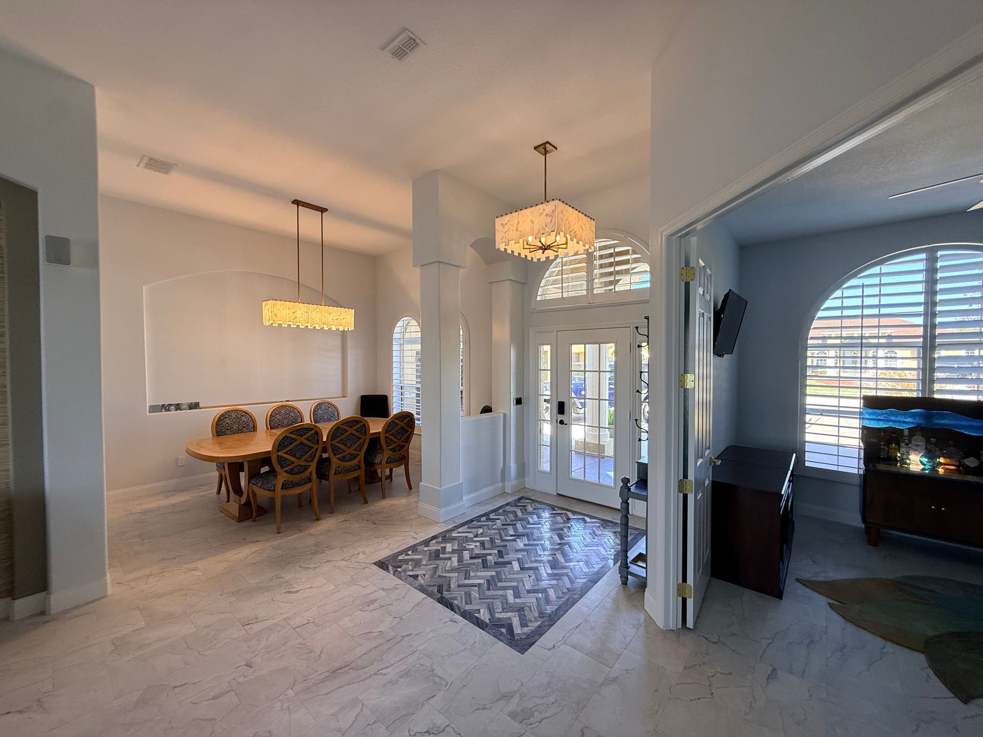 A bright foyer and dining area with light tile floors, a patterned rug, two modern chandeliers, and arched windows.