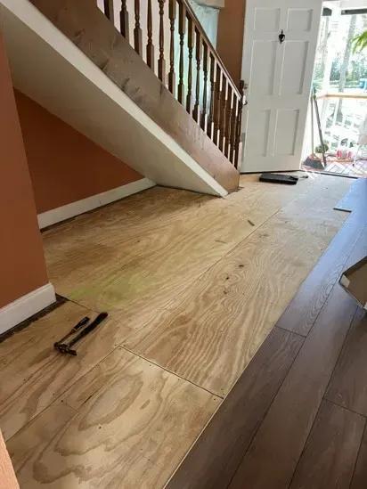 Plywood subfloor under staircase in a hallway, with tools on the floor and light from a doorway.