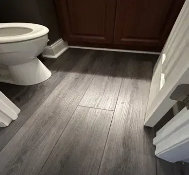 Small bathroom with dark wood-look floor tiles, toilet on the left, and cabinet doors ahead.