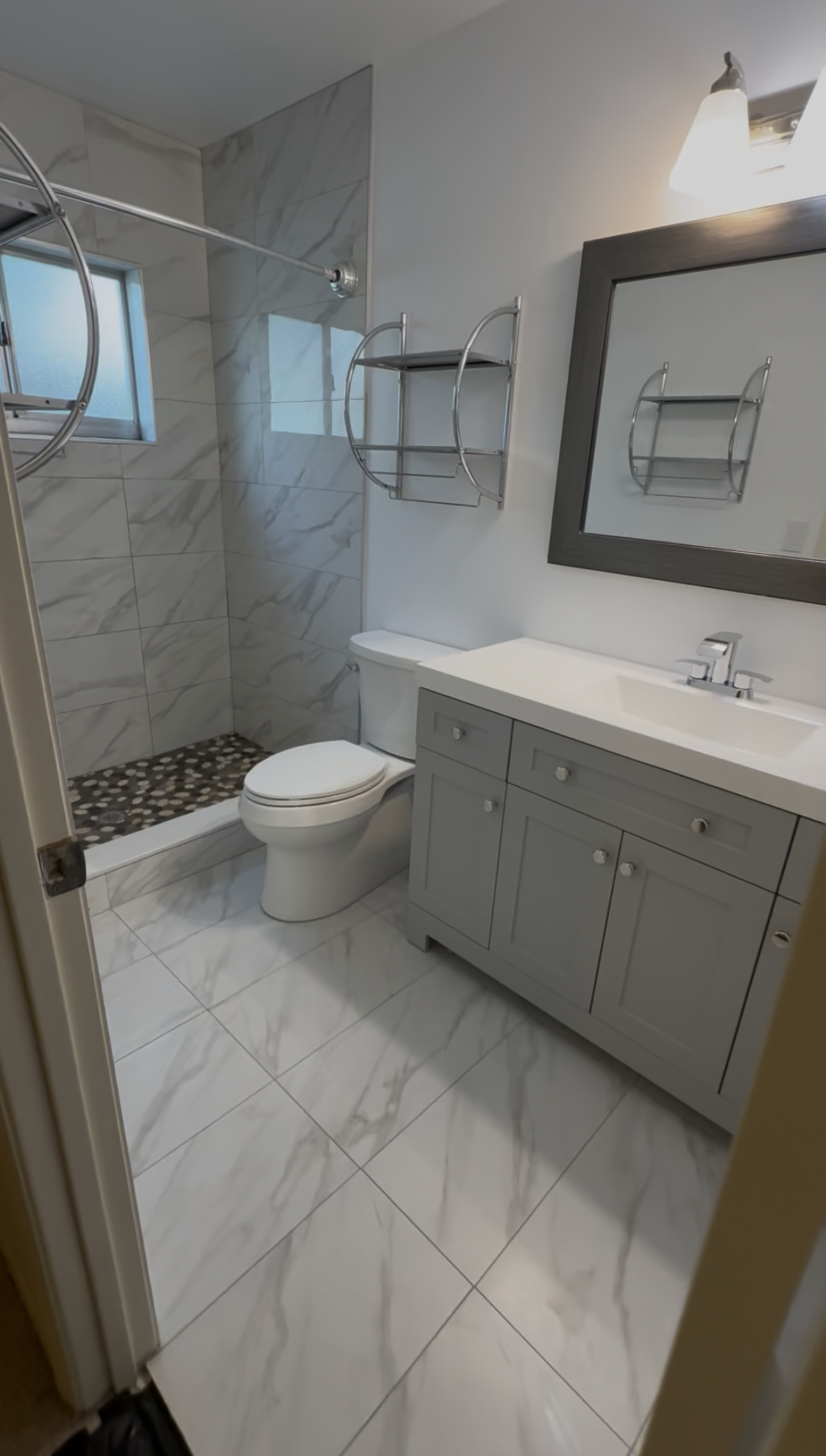 Modern bathroom with white tile, gray vanity, toilet, wall mirror, and glass shower.