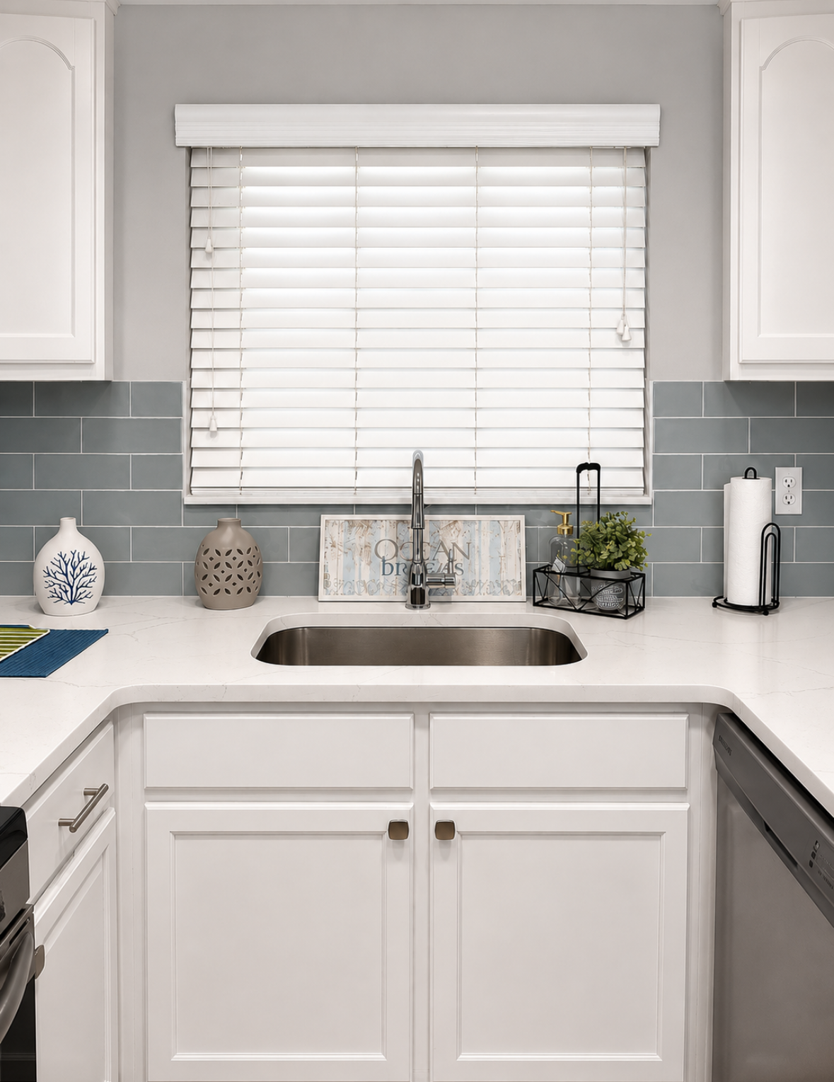 Bright white kitchen sink with window blinds, gray tile backsplash, and white cabinets.