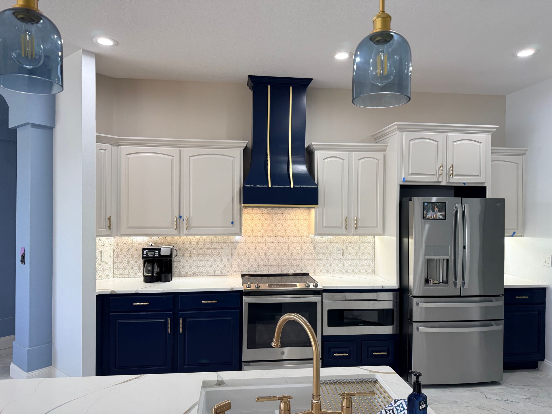 A kitchen with white upper cabinets, dark blue lower cabinets, a blue range hood, stainless appliances, and pendant lights.