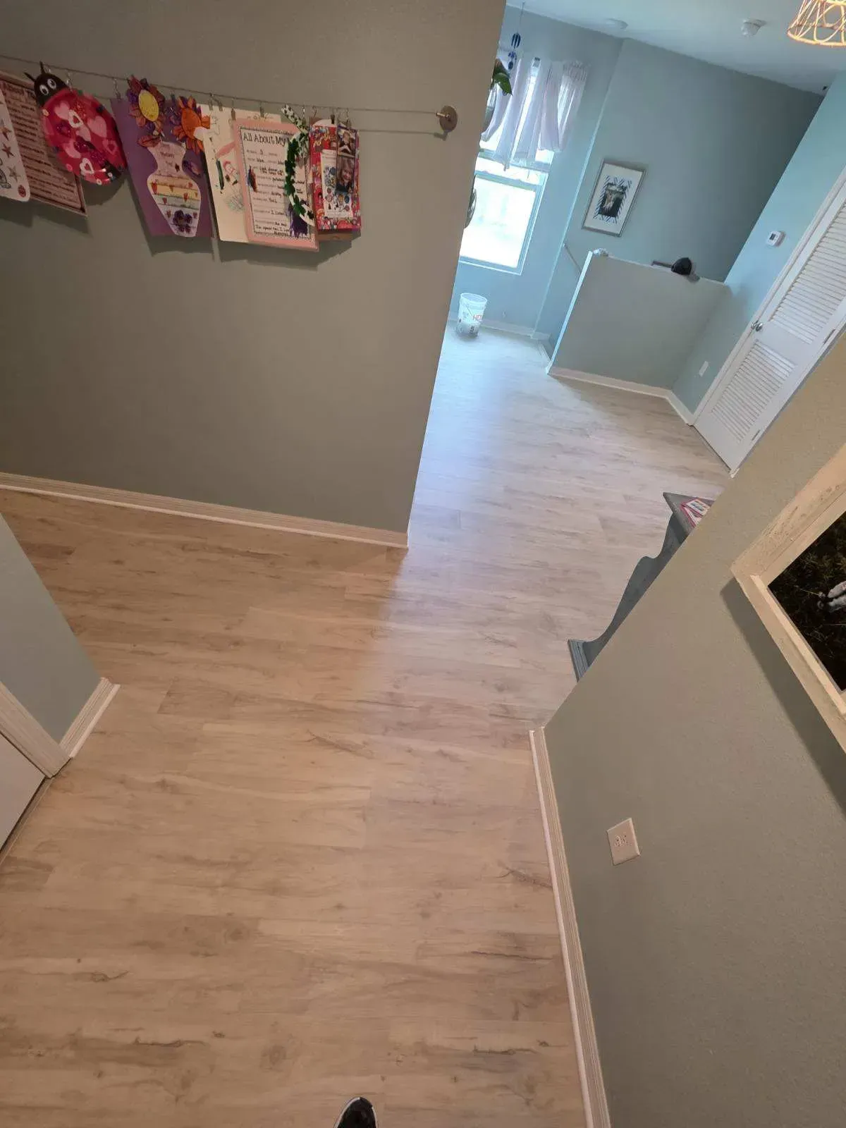 Hallway with light wood floors, gray walls, and children’s artwork hanging near a bright doorway