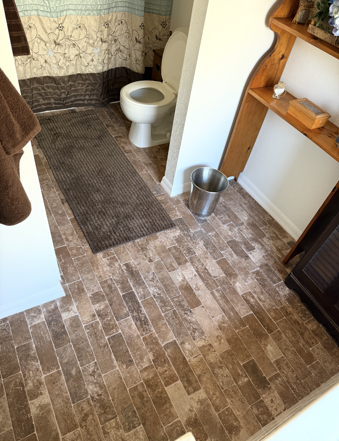 Small bathroom with a white toilet, dark bath mat, tiled floor, and a metal bucket near a wooden shelf.