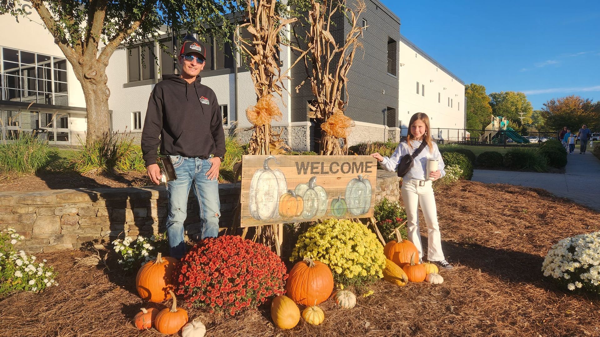 Two people stand by a "WELCOME" sign decorated with pumpkins and fall flowers outside a building.