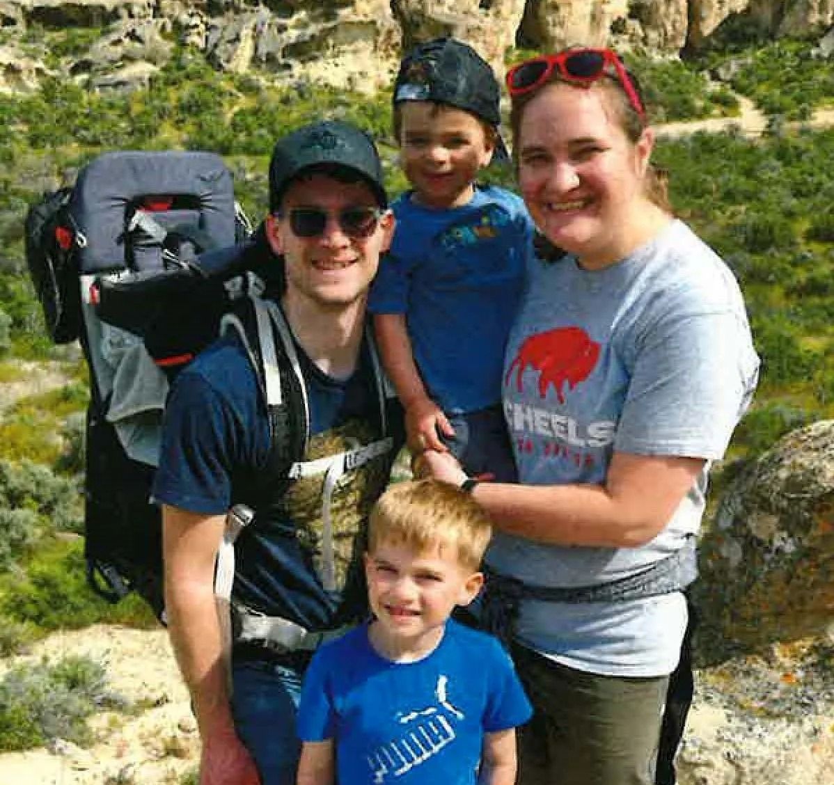 Family of four smiling outdoors near rock formations; man with a child carrier, boy, woman holding another boy.
