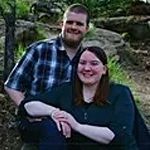 Couple smiles in outdoor setting; man in plaid shirt, woman in teal top.