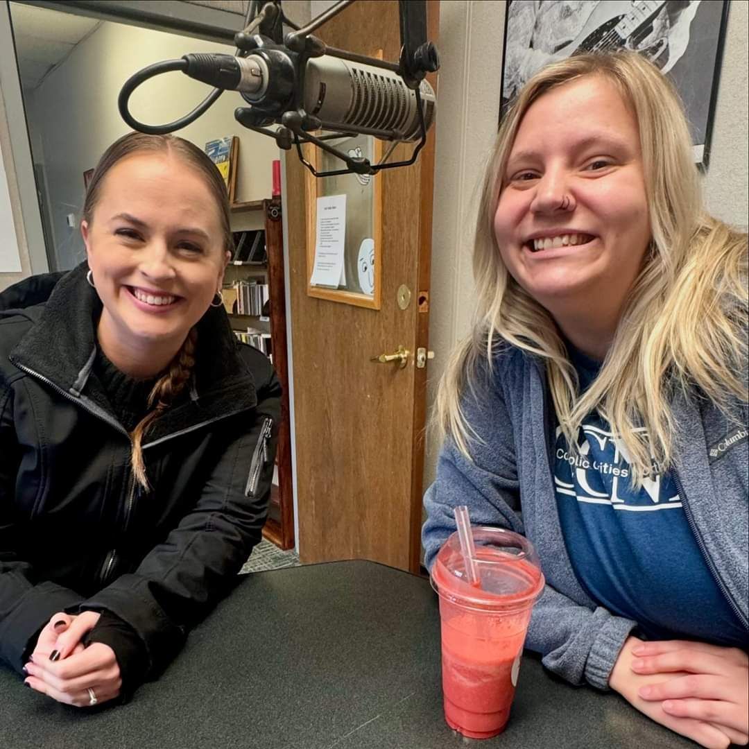 Two women smiling at a table in a studio, facing a microphone. One holds a pink drink.