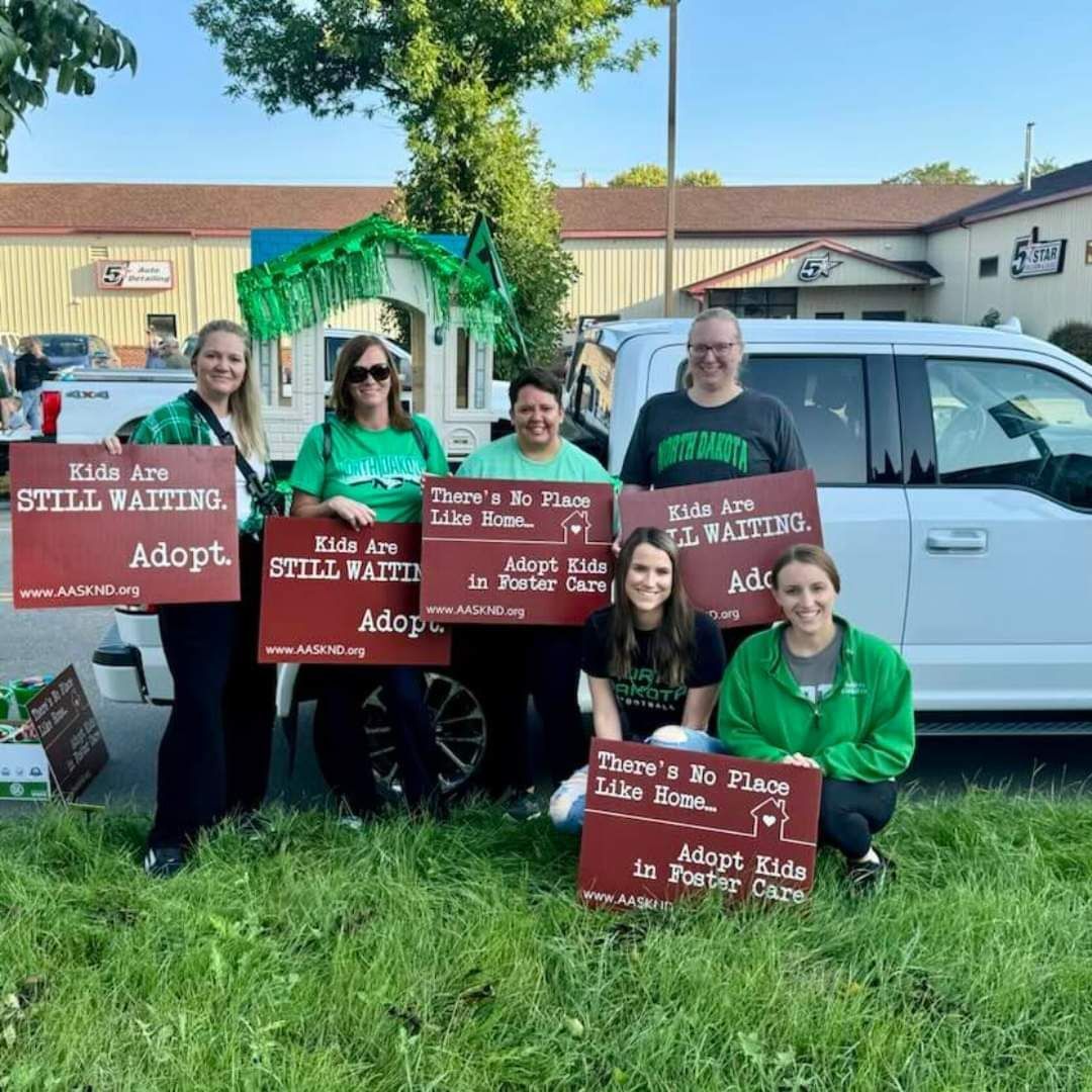 People holding signs about adopting kids pose in front of a truck, near a building.