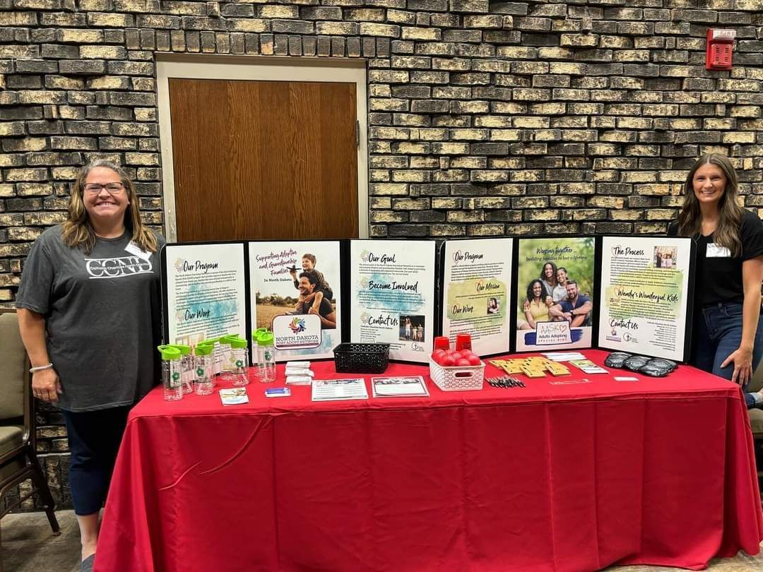 Two women stand behind a table displaying posters and materials. The table is red. Brick wall backdrop.