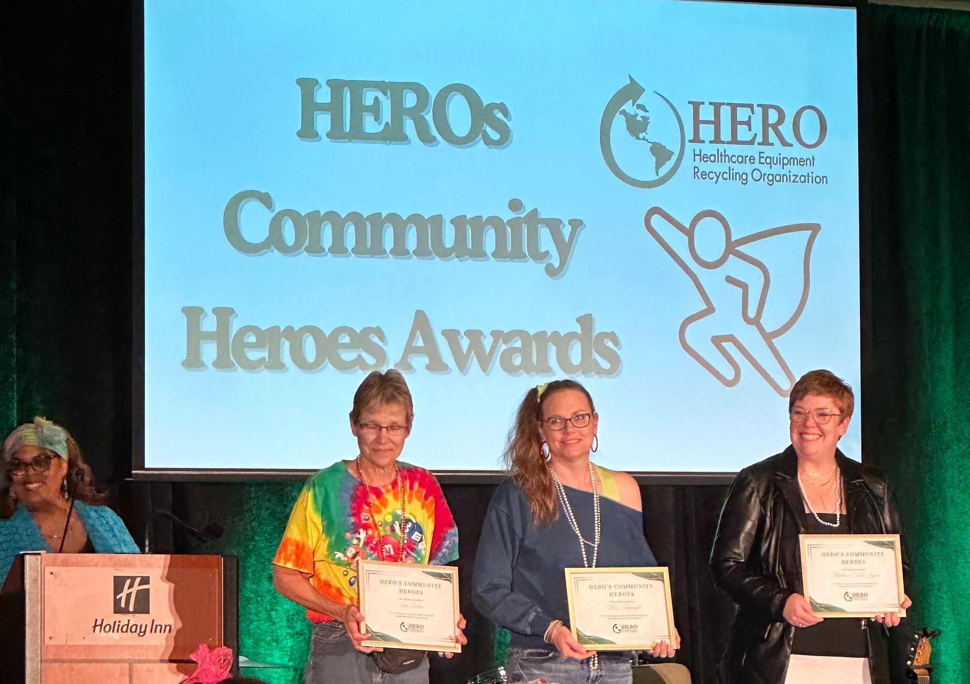 HEROs Community Heroes Awards ceremony. Three people hold certificates on a stage with a screen and logo.