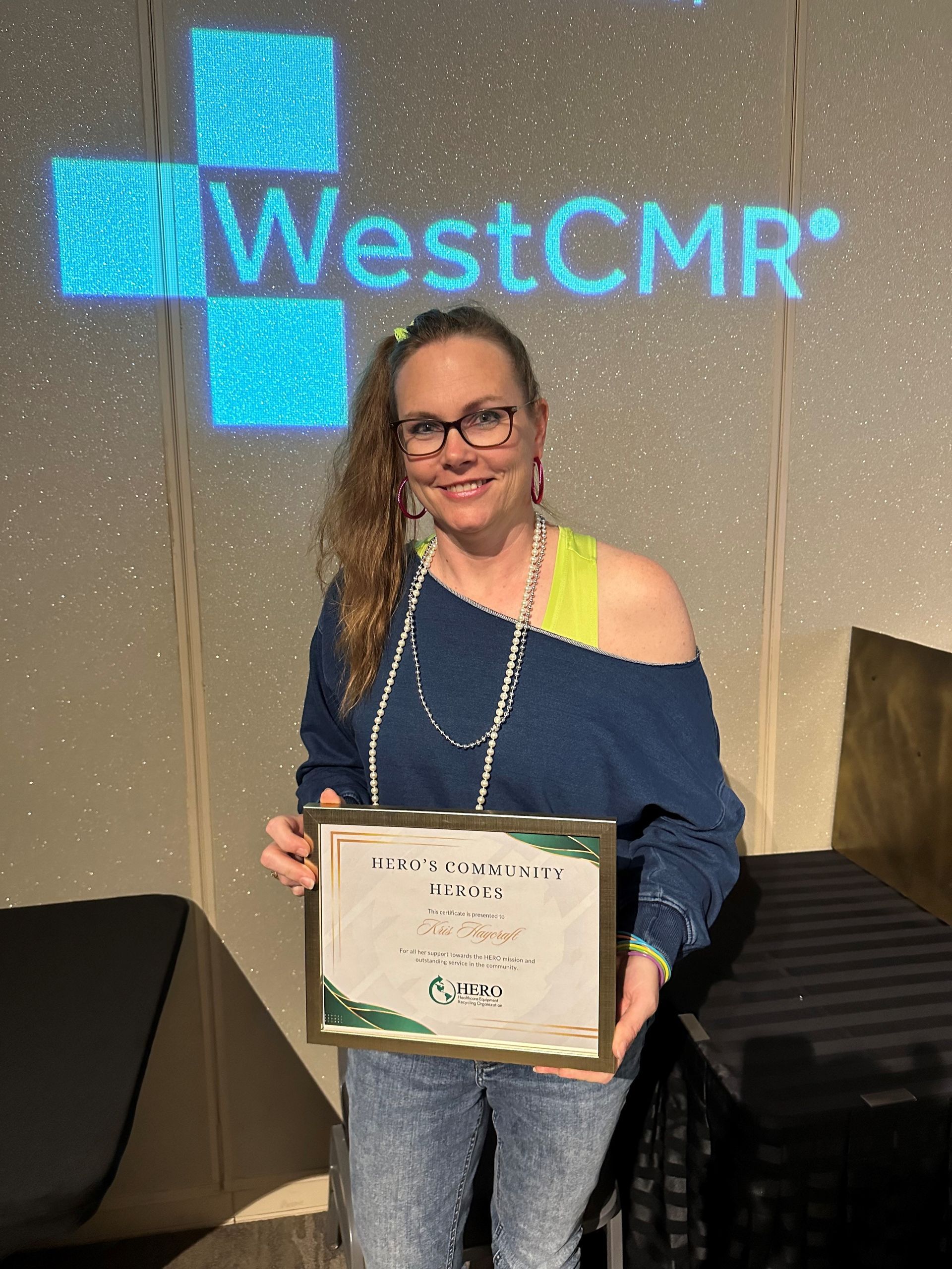 Woman holding an award in front of a WestCMR logo; she wears a blue top, jeans, and glasses.