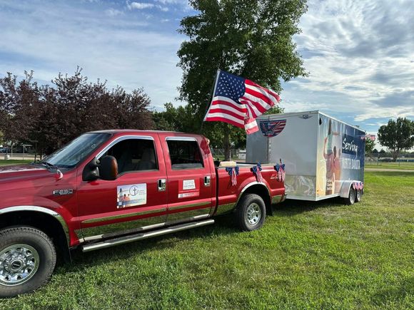Red pickup truck towing a trailer, both decorated with American flags, parked on grass.